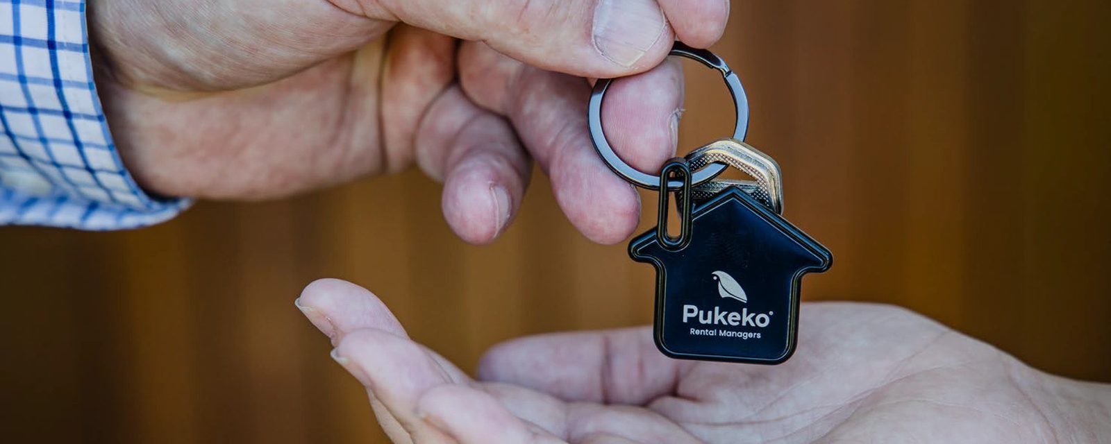 A hand in a checked shirt cuff holding out a Pukeko house-shaped keyring on a wooden background.